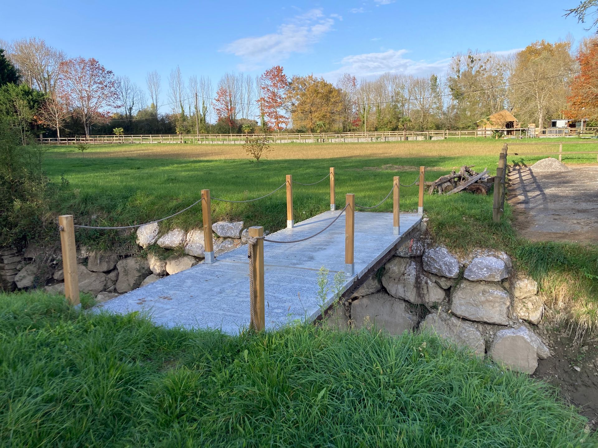 Pont de pierres et de gravier enjambant un petit fossé dans un champ herbeux. Poteaux en bois et rambarde en chaînes.