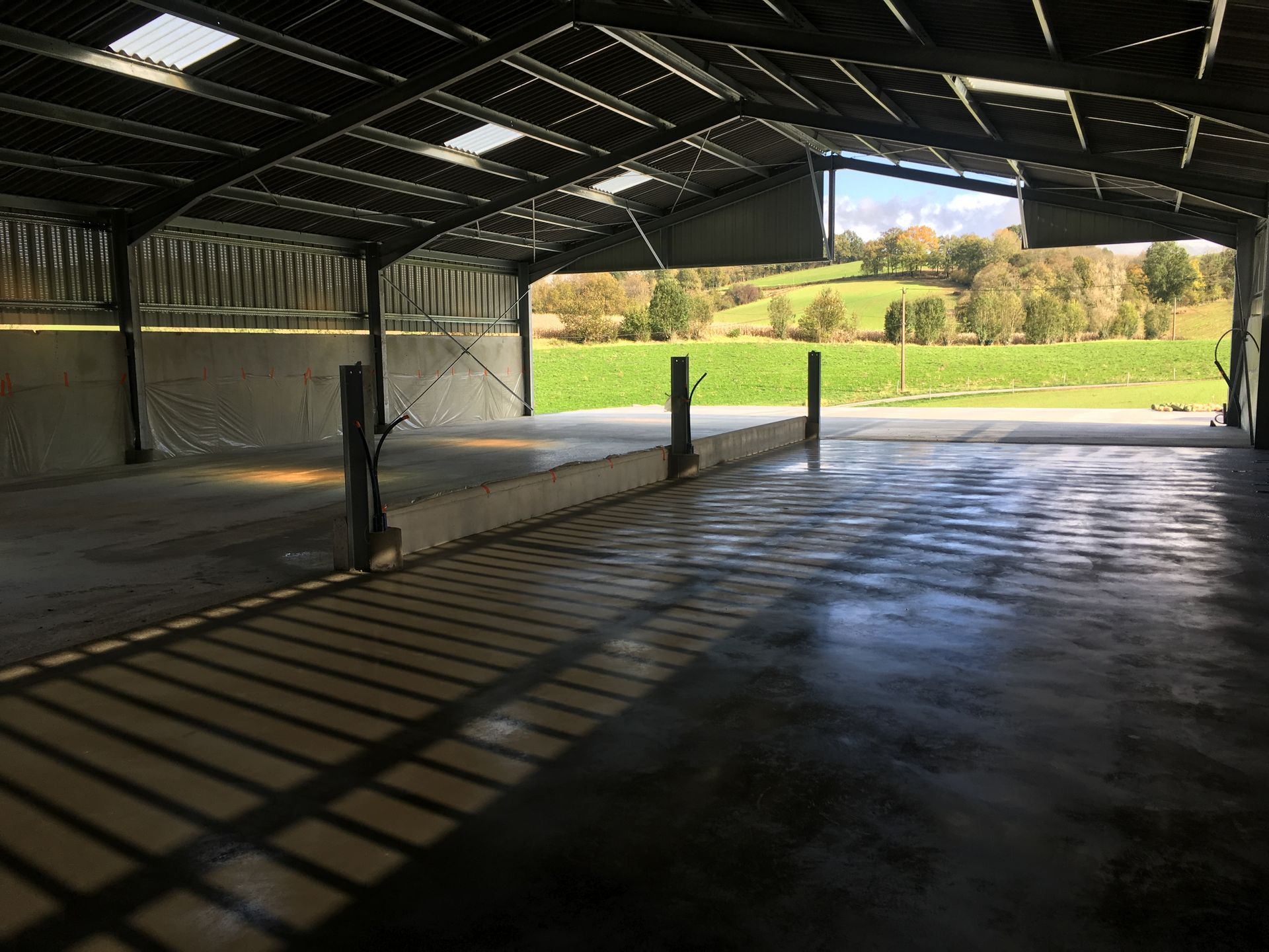 Intérieur d'une grange métallique vide, avec sol en béton et vue sur des champs verdoyants et des collines.