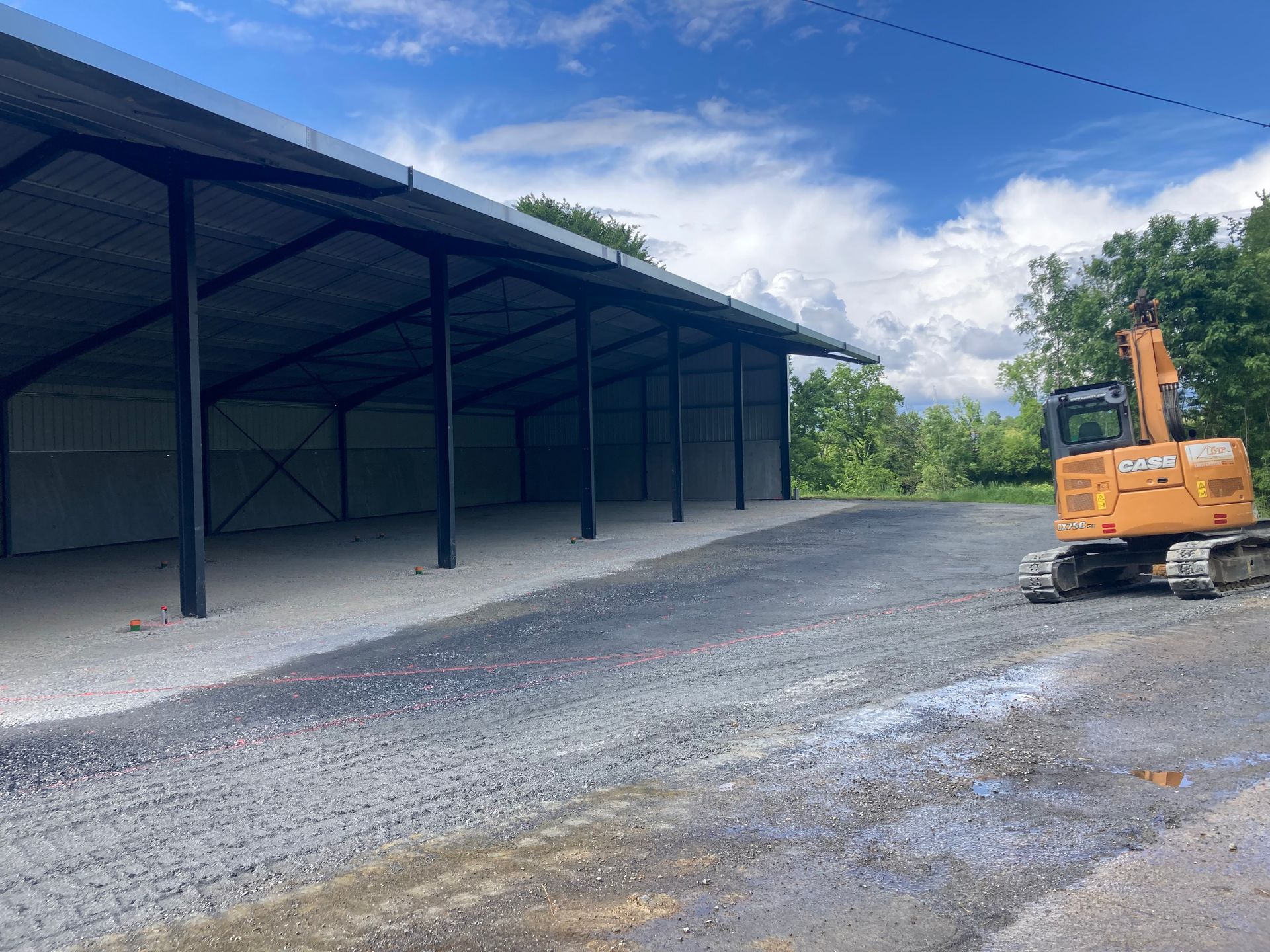 Une allée de gravier mène à un hangar métallique ouvert sur un côté, avec une excavatrice sur la droite.