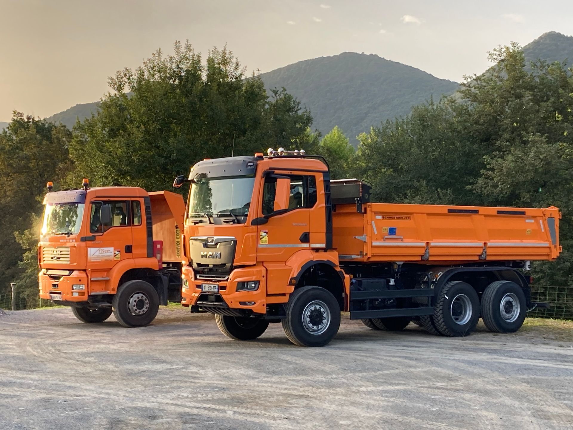 Deux camions-bennes orange stationnés à l'extérieur, avec une montagne en arrière-plan.