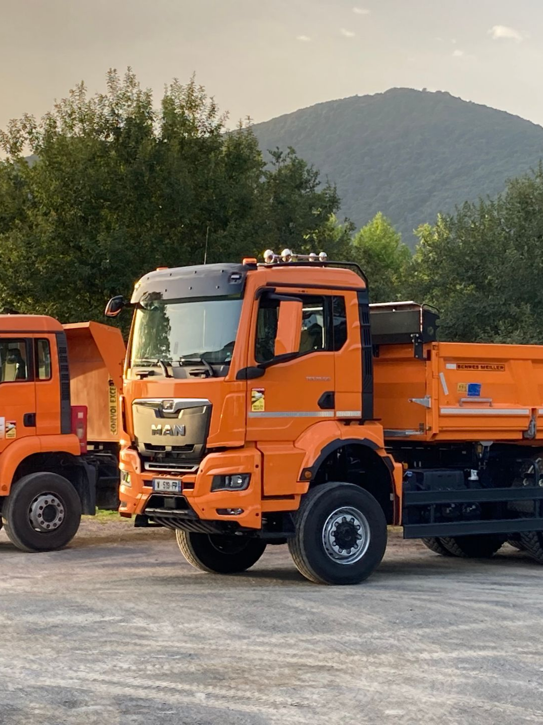 Deux camions-bennes orange sont stationnés à l'extérieur, près d'arbres et d'une montagne.