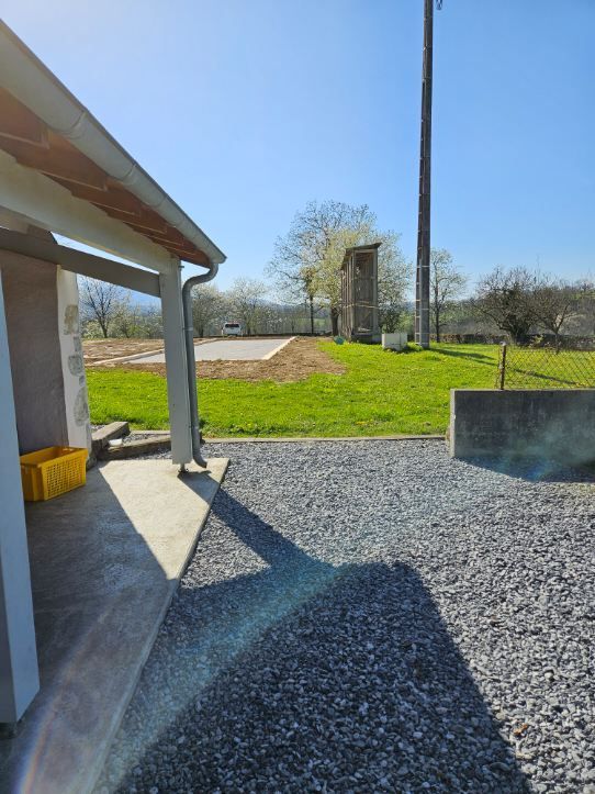 Un bâtiment avec un porche, un sol gravillonné et un champ verdoyant en arrière-plan, par une journée ensoleillée.