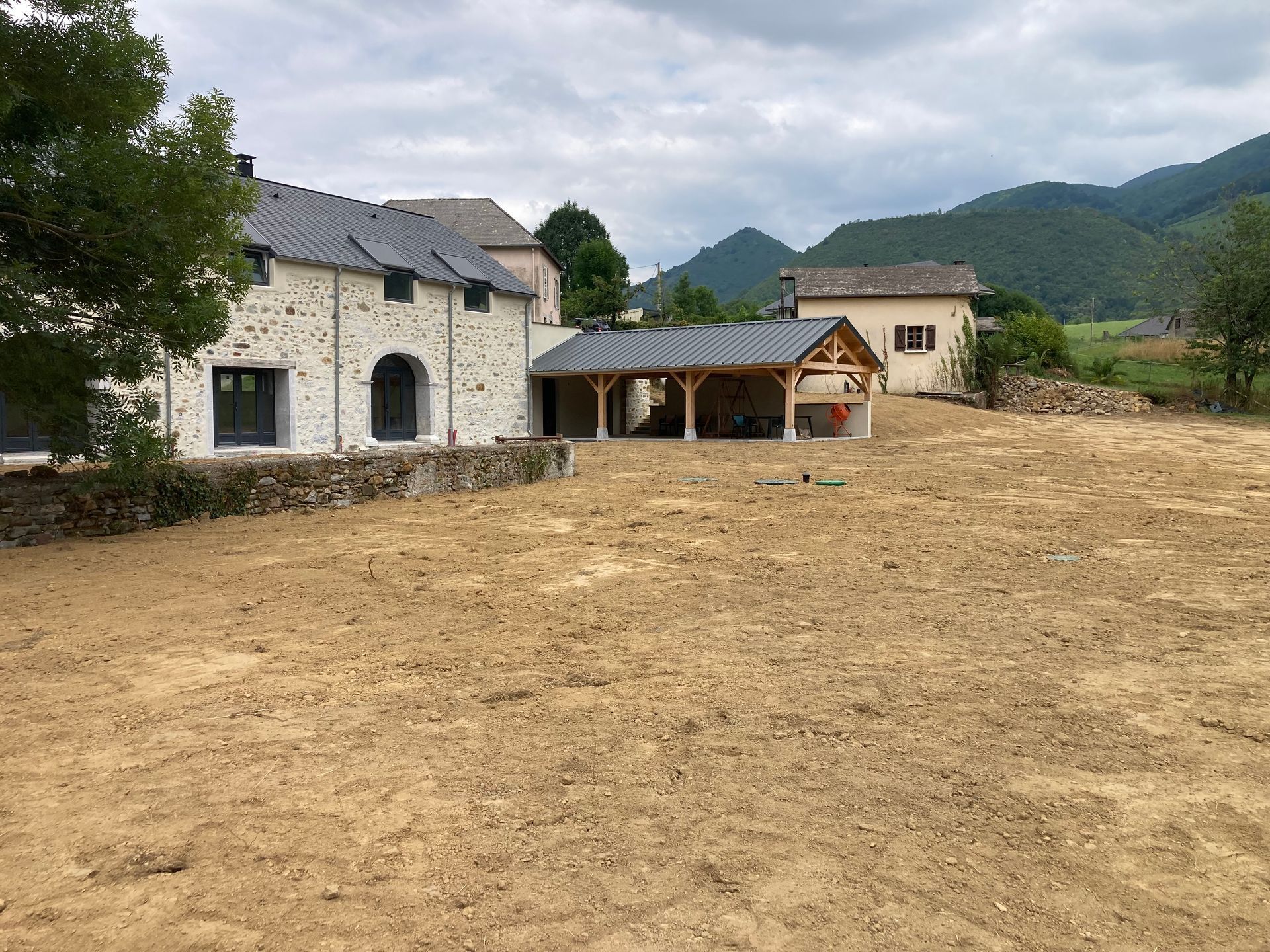 Une scène rurale avec des bâtiments en pierre, un abri en bois et des montagnes en arrière-plan, sous un ciel nuageux.