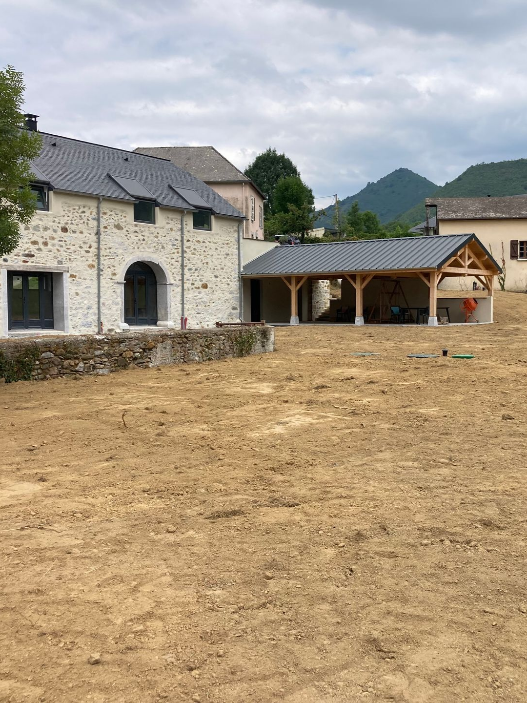 Bâtiment en pierre avec une structure en bois couverte et une cour en terre battue devant. Montagnes en arrière-plan.