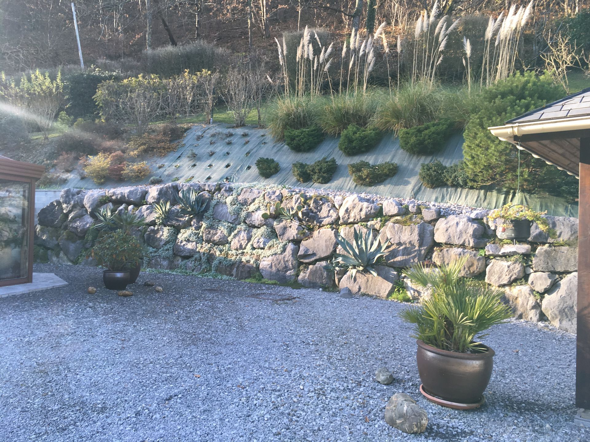 Mur de soutènement en pierre avec plantes, terrasse en gravier et jardin à flanc de colline sous un ciel ensoleillé.