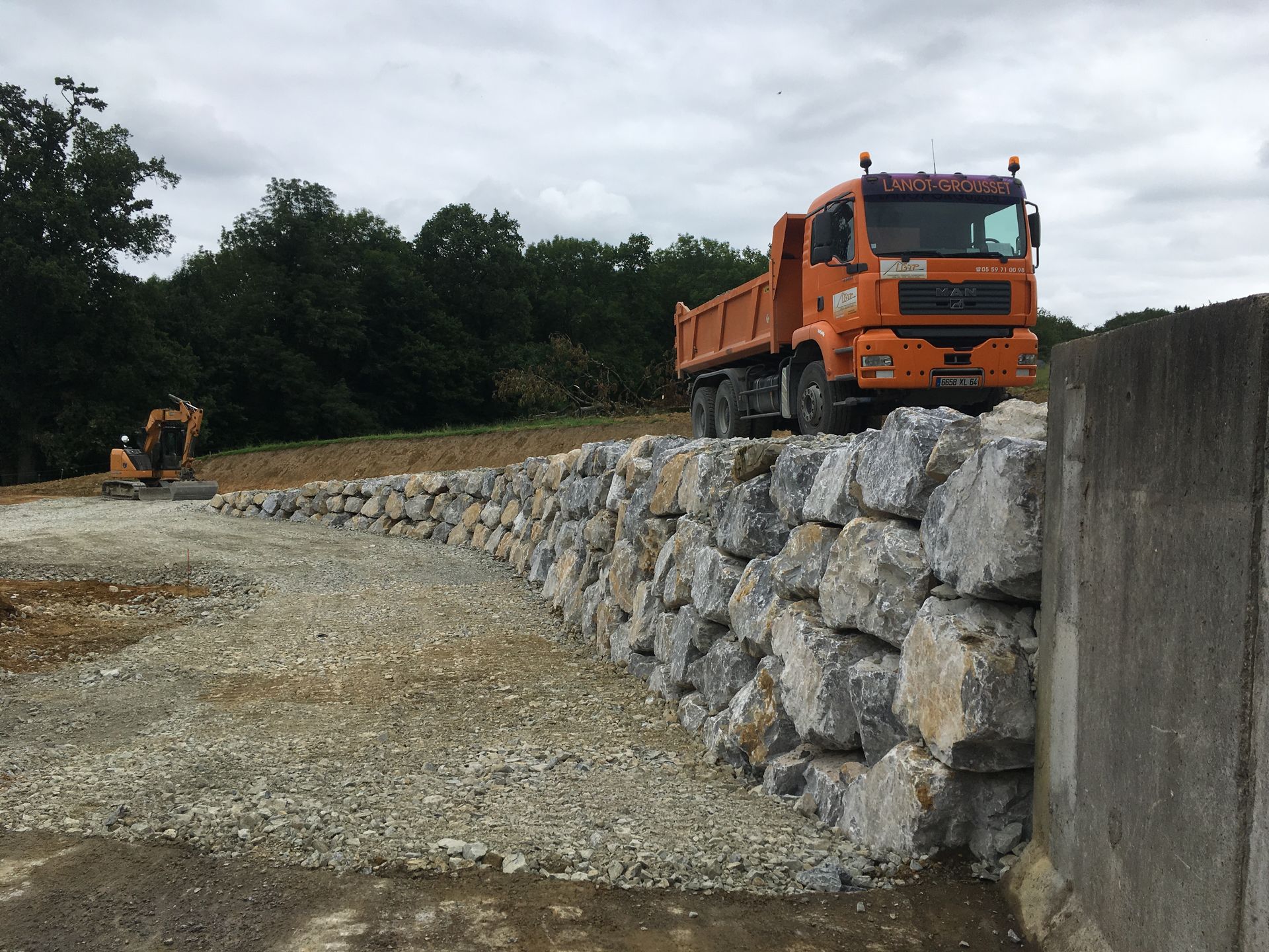 Chantier : camion-benne orange sur un mur de roche, excavatrice en arrière-plan.