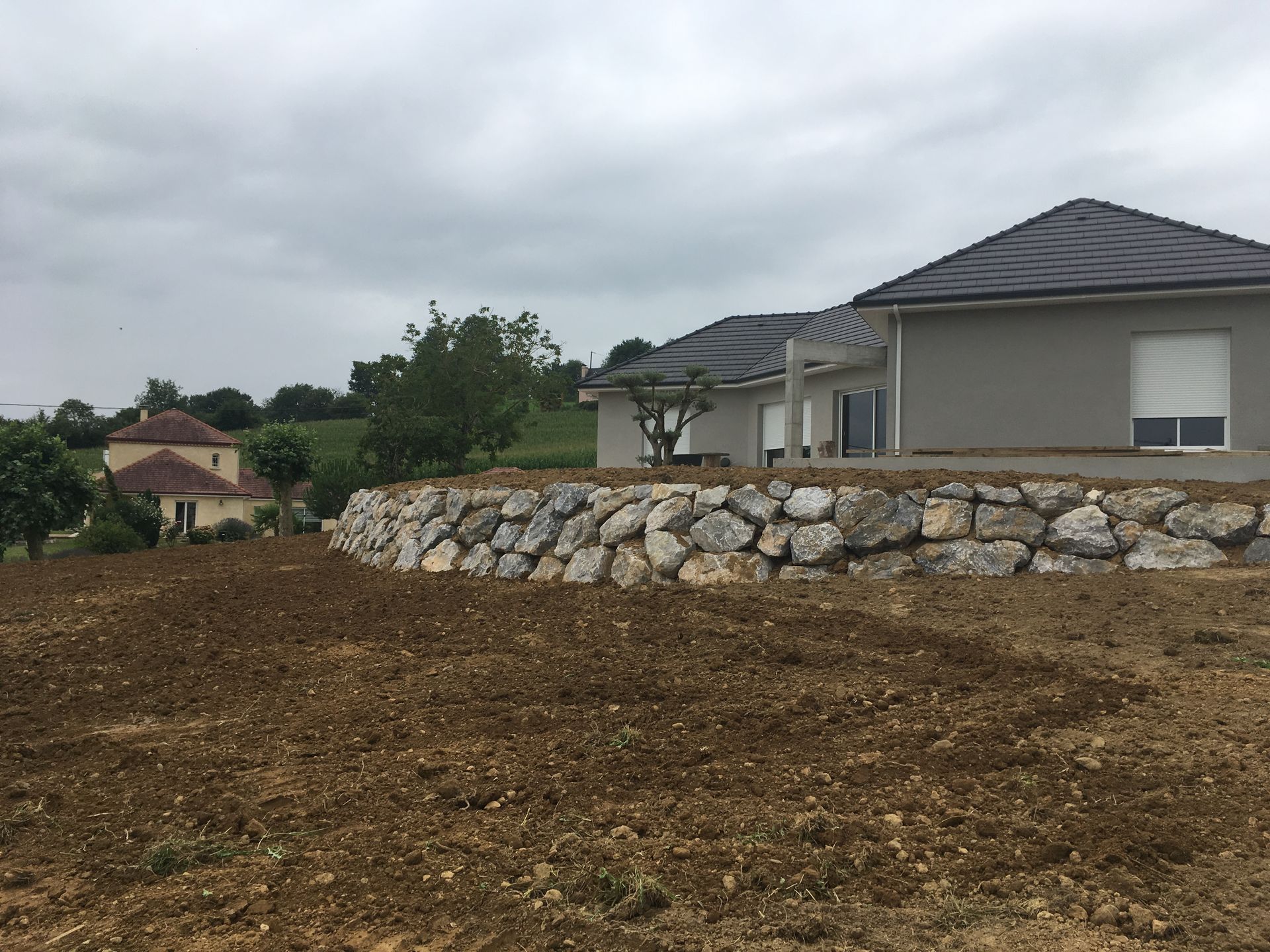 Une maison moderne avec un mur de soutènement en pierre et un ciel nuageux dans un cadre champêtre.