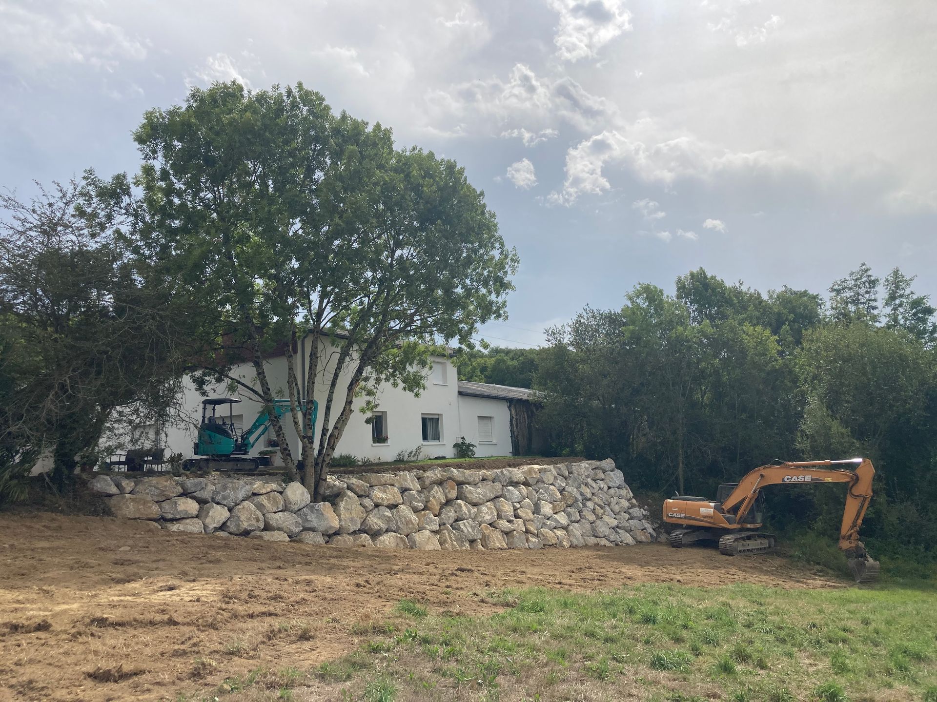 Chantier de construction avec une maison blanche, un mur en pierre et deux excavatrices.