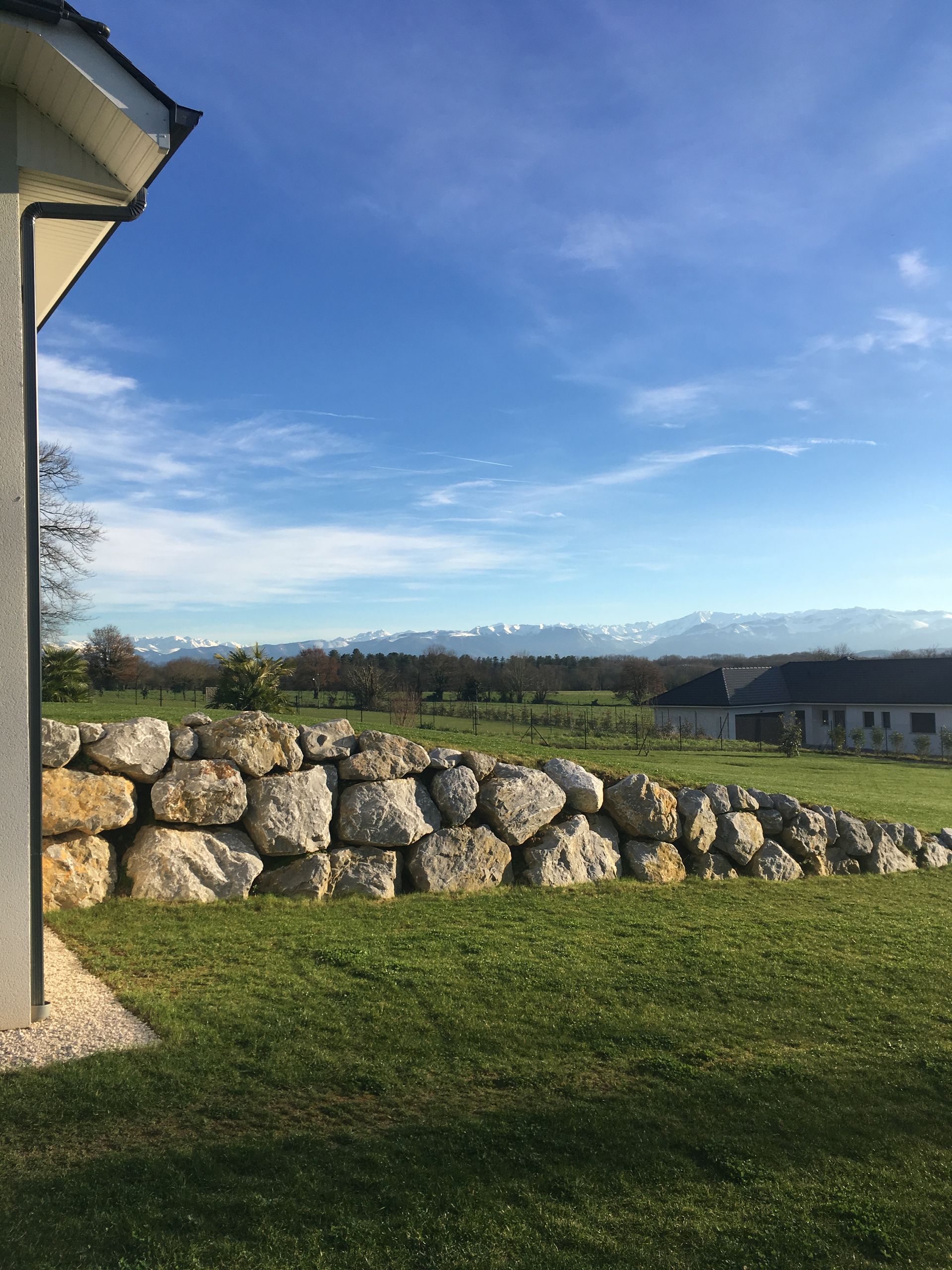 Un mur de pierre dans un champ herbeux, avec une maison et des montagnes en arrière-plan, sous un ciel bleu.