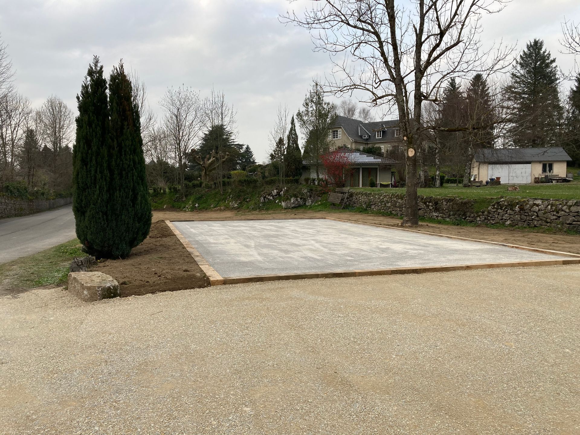 Terrain gravillonné avec une place pavée, un grand conifère et des maisons en arrière-plan.