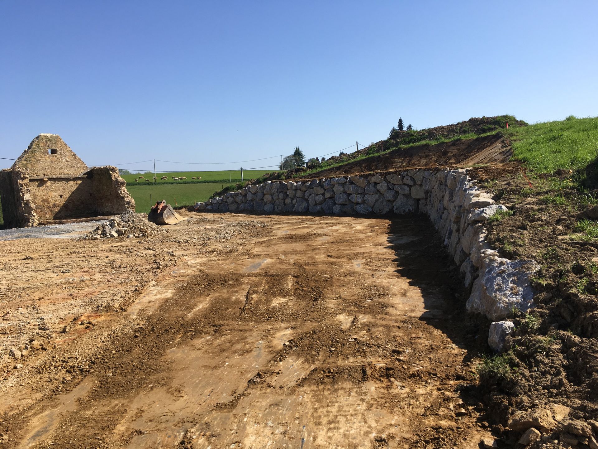 Chantier avec mur de soutènement en pierre. Un chemin de terre et un petit bâtiment en ruine sont visibles.