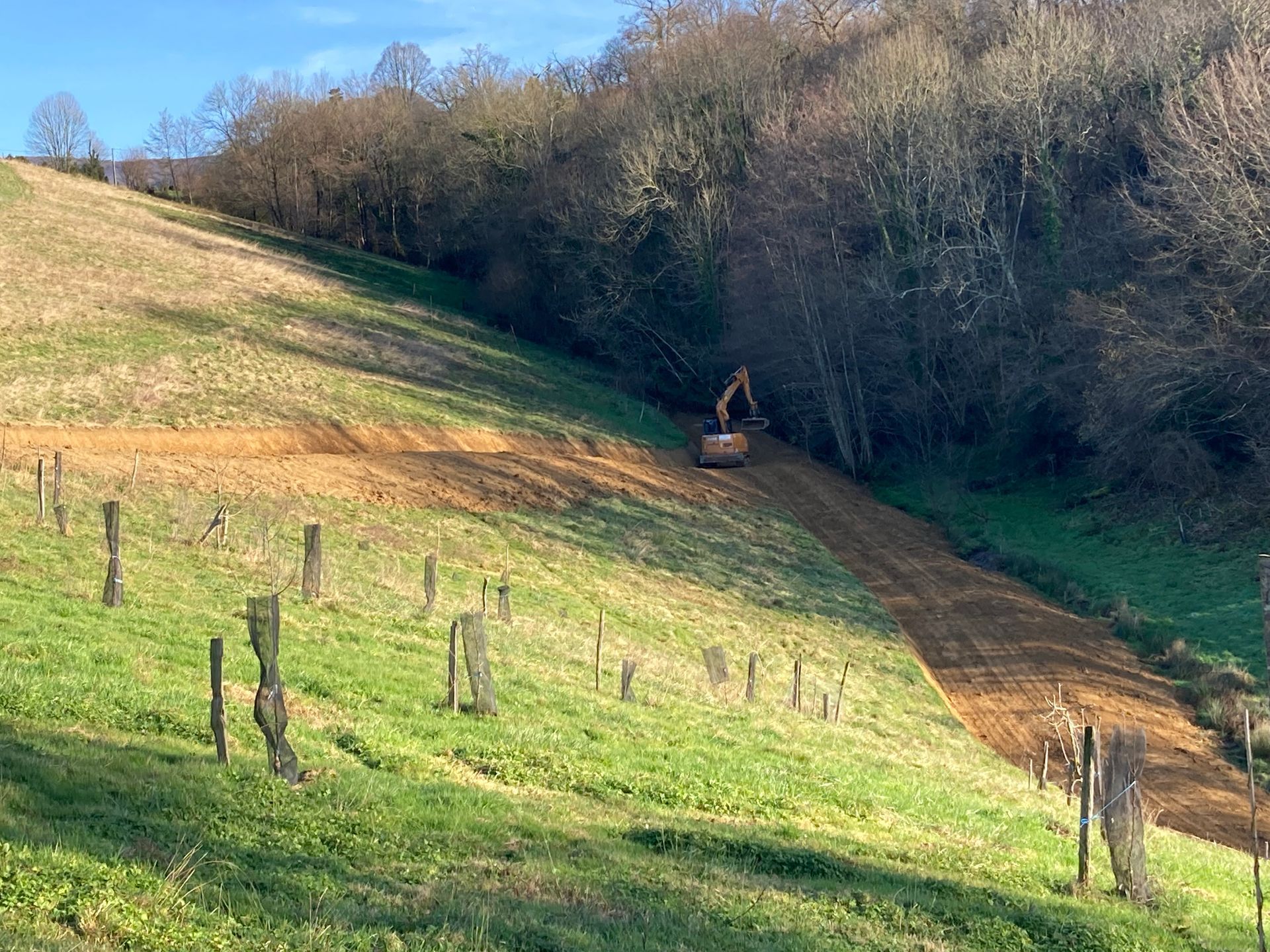 Une pelleteuse travaille à flanc de colline, dégageant un chemin. La terre brune contraste avec l'herbe verte et les arbres.
