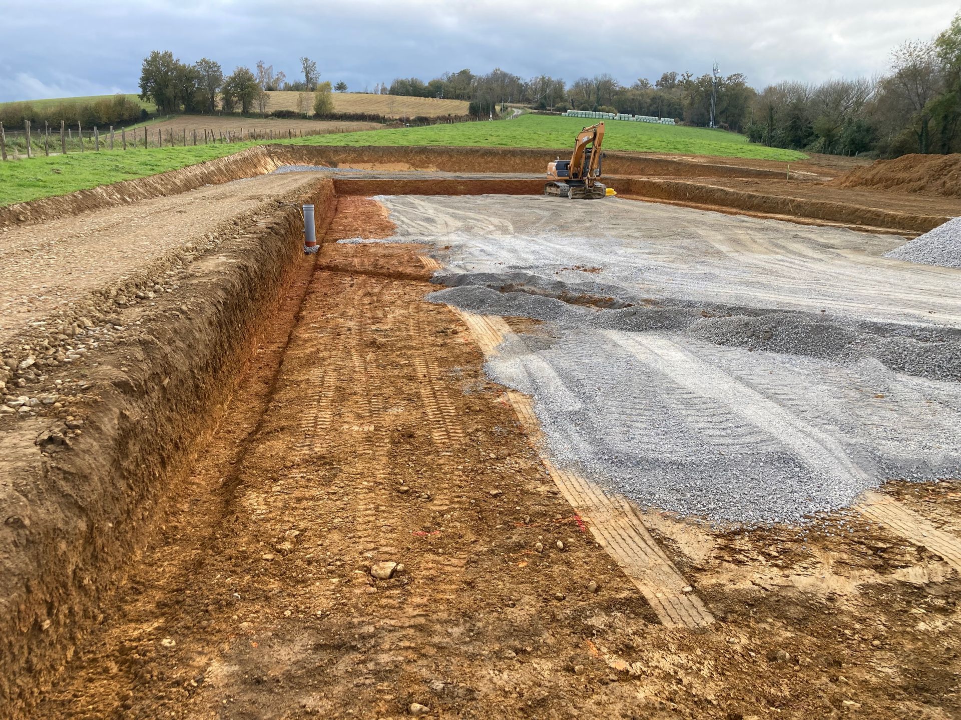 Chantier : terre excavée, base de gravier, pelle mécanique, cadre rural, ciel couvert.