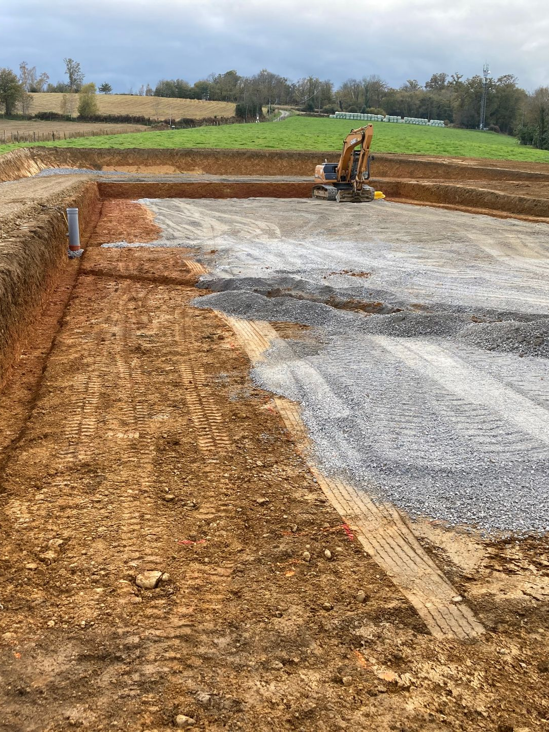 Chantier de fondations avec une excavatrice. Des murs de terre entourent une base de gravier et de terre.