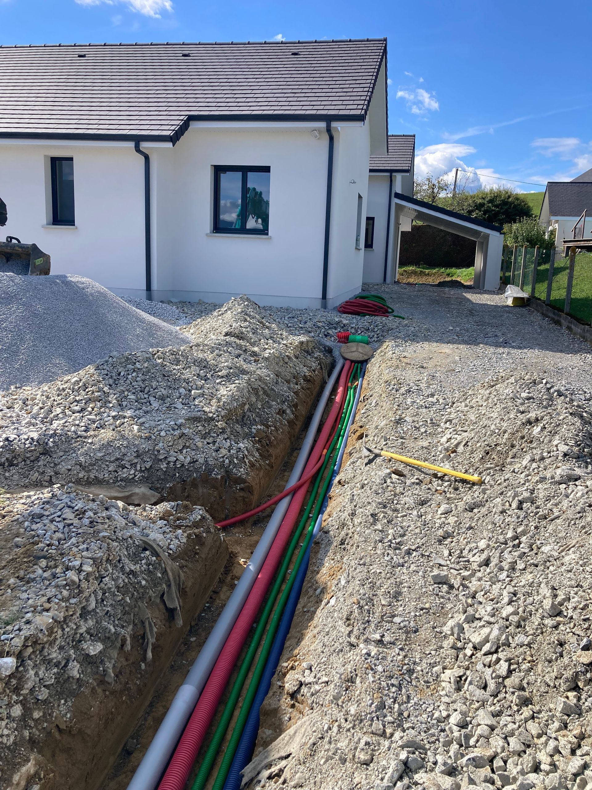 Une tranchée bordée de conduits colorés mène à une maison blanche au toit brun ; le sol est recouvert de gravier.