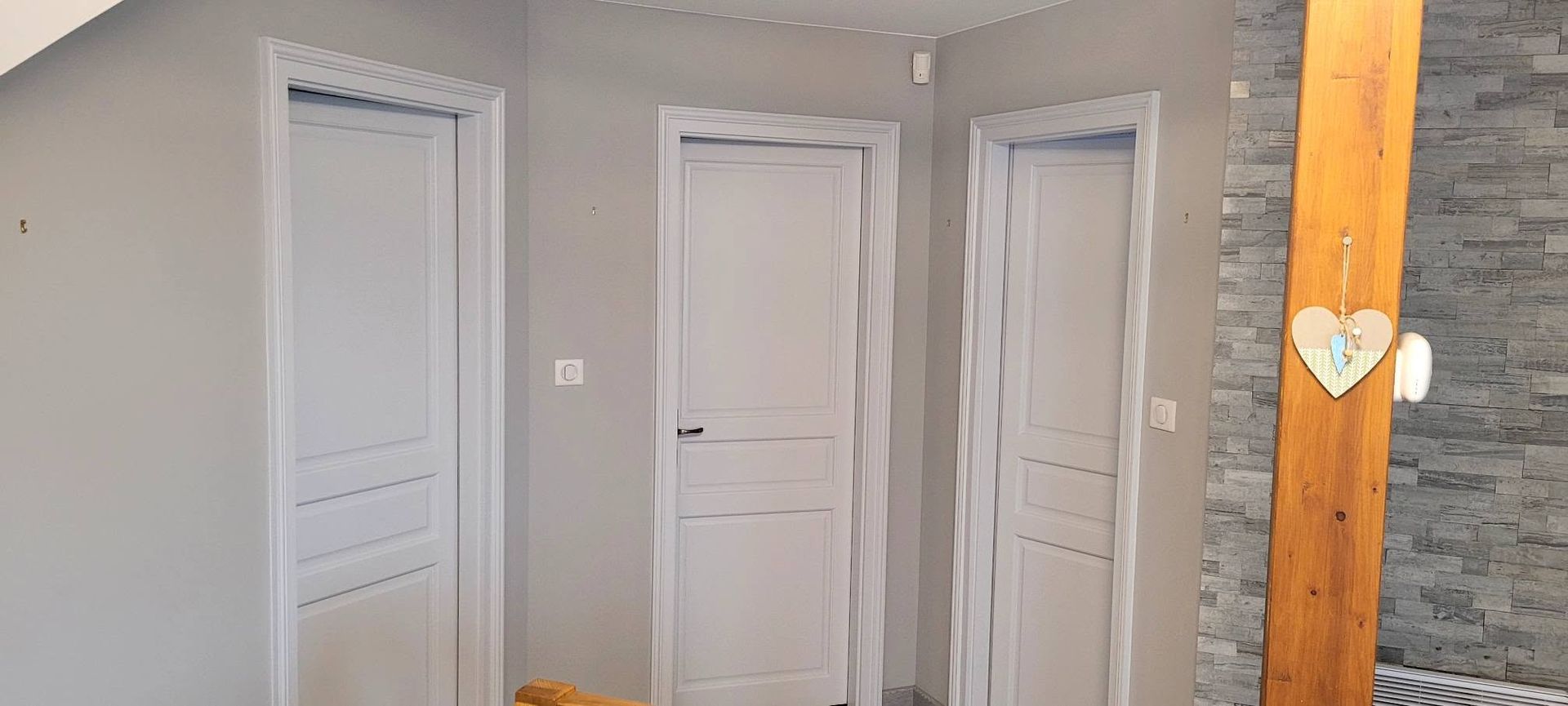Trois portes intérieures blanches dans un couloir gris, de part et d'autre d'un poteau en bois et d'un mur en pierre.