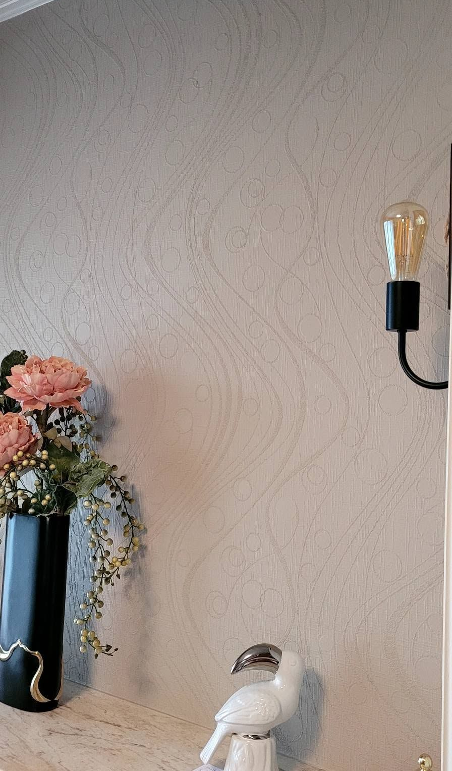 Vase de fleurs roses et figurine d'oiseau blanc sur une étagère à côté d'une applique murale sur un mur beige