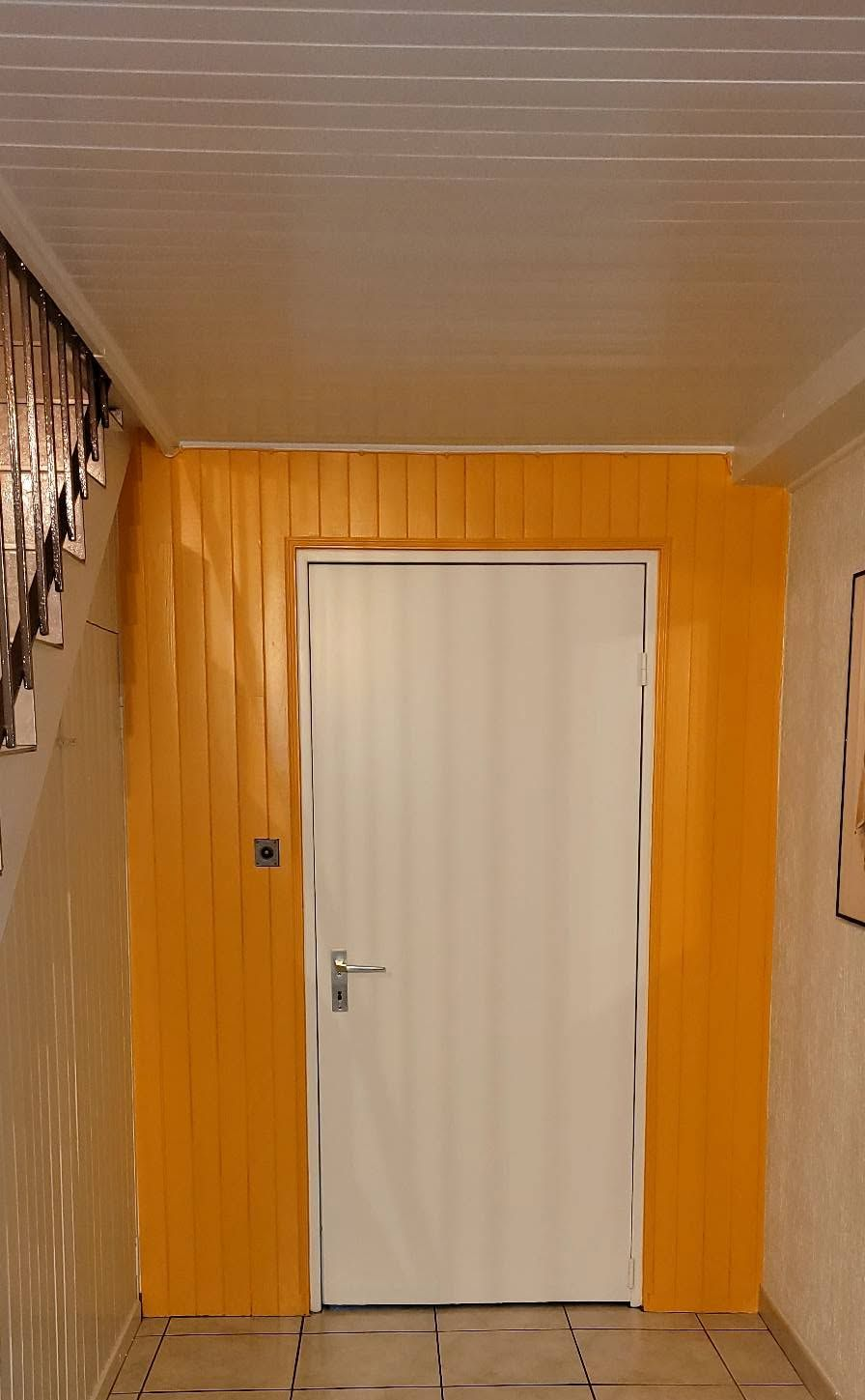 Couloir étroit avec une porte blanche centrée dans un mur lambrissé orange et un sol carrelé
