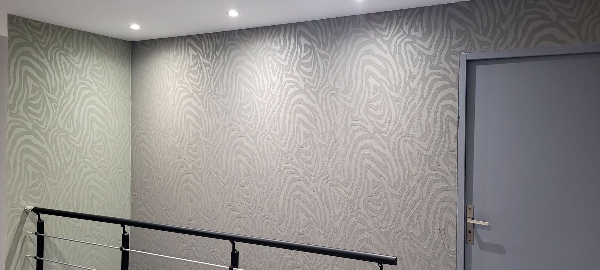Couloir avec papier peint à motifs, rampe noire et porte grise sous éclairage encastré au plafond