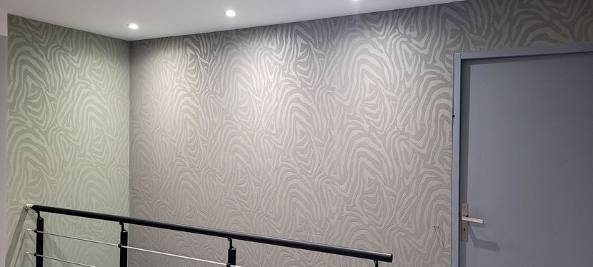 Couloir avec papier peint à motifs, rampe noire et porte grise fermée sous des spots encastrés au plafond