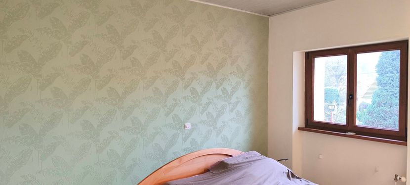 Chambre vide aux murs vert pâle, avec une fenêtre et un lit défait au premier plan.
