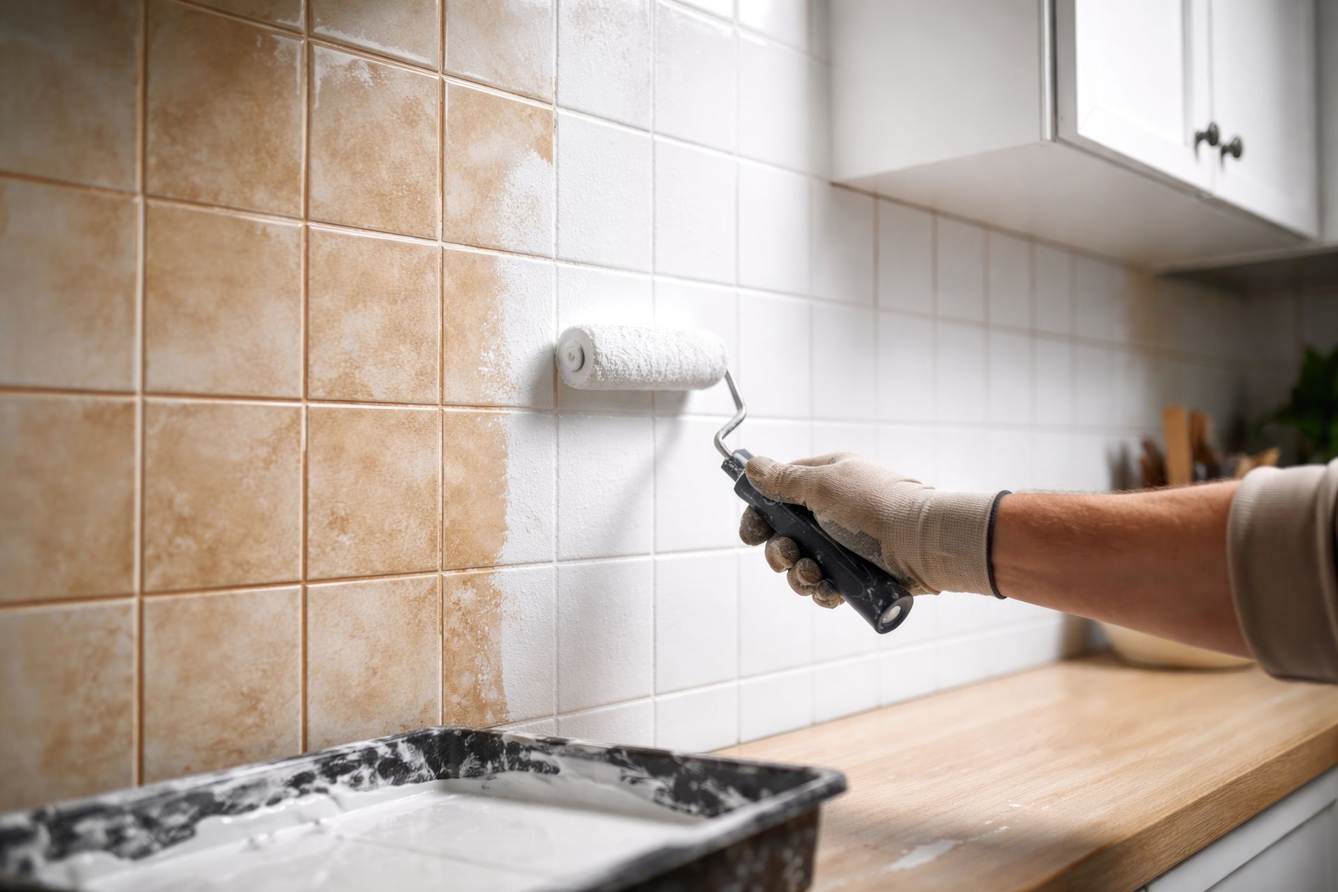 Main gantée peignant au rouleau un mur beige de cuisine près du plan de travail et de la crédence carrelée