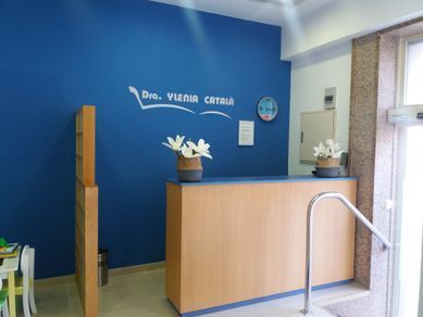 Zona de recepción con pared azul, mostrador de madera, nombre “Dra. Vlenia Catala” y una pequeña planta.