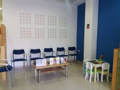 Sala de espera con sillas azules, una mesa pequeña con folletos y una mesa para niños. Paredes blancas y azules.