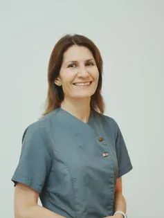 Mujer de cabello castaño, sonriente, vistiendo una parte superior de uniforme verde azulado, sobre un fondo blanco.