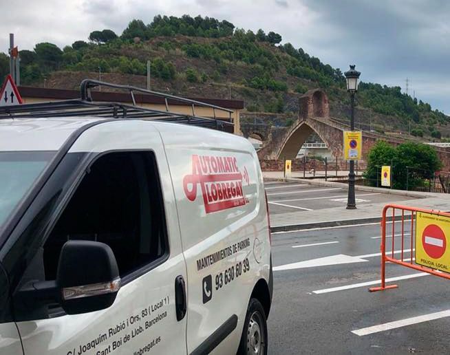 Furgoneta blanca con logotipo rojo estacionada en la calle, con un puente arqueado medieval al fondo.