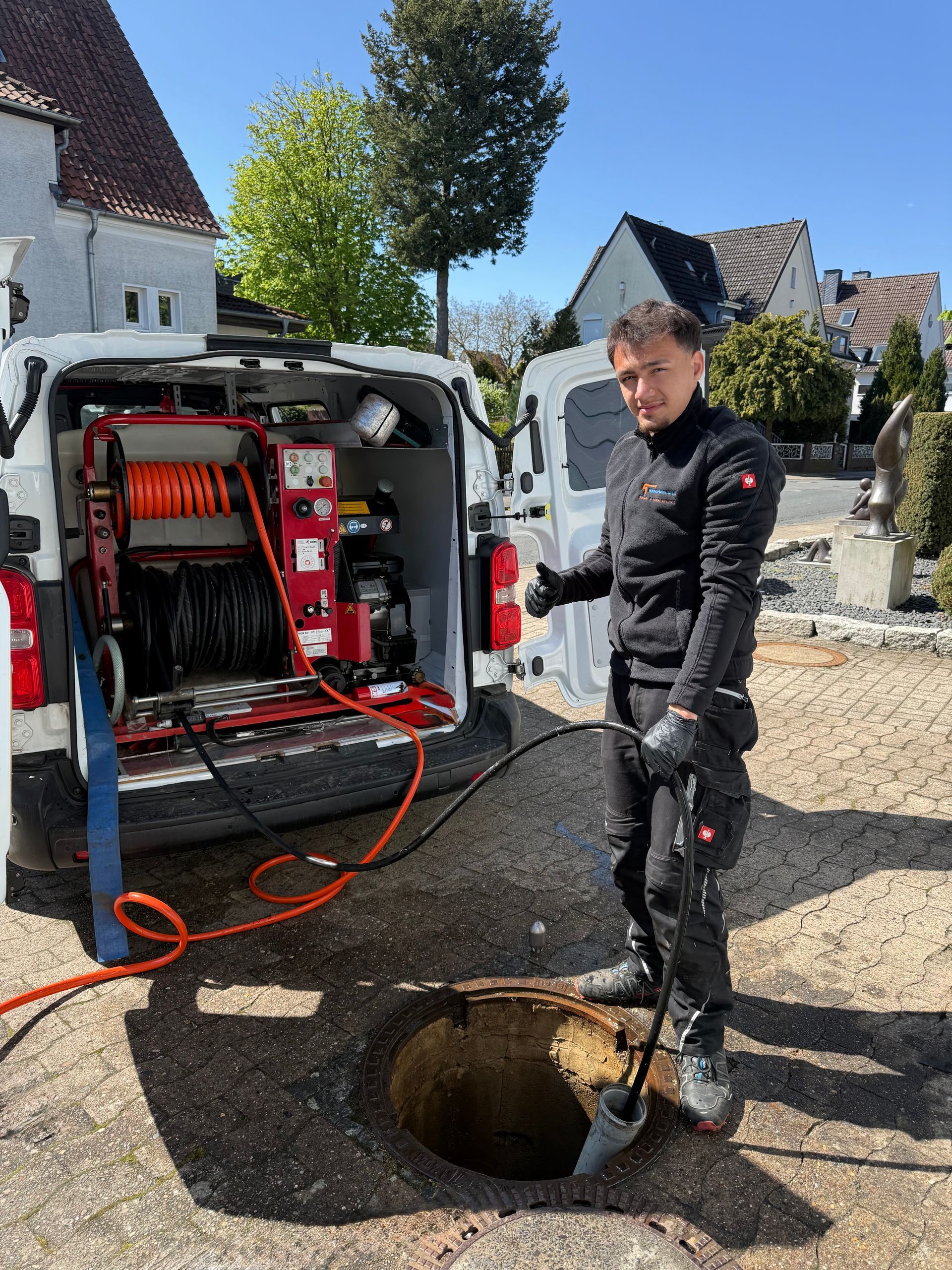 Ein Arbeiter in schwarzer Arbeitskleidung steht neben einem offenen Lieferwagen und einem Kanaldeckel in einer Wohnstraße.