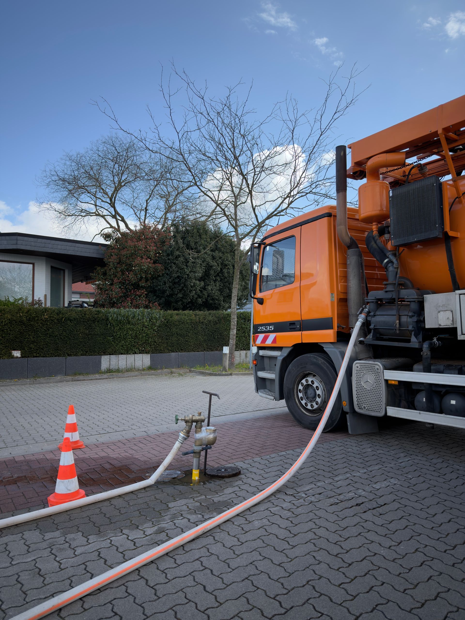Ein orangefarbenes Versorgungsfahrzeug ist neben Verkehrskegeln in einer Wohnstraße an einen Schlauch angeschlossen.