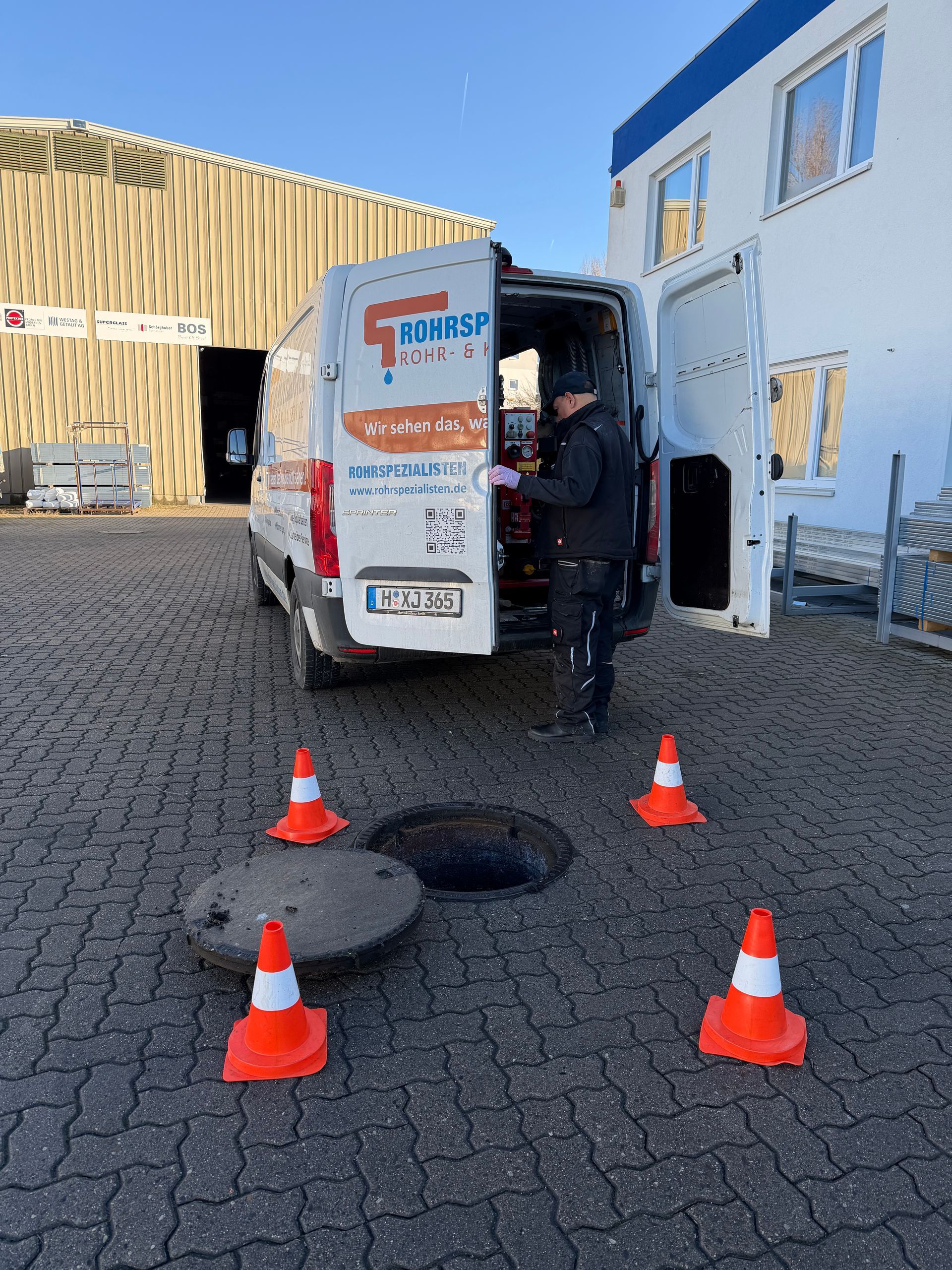 Arbeiter entlädt einen Lieferwagen in der Nähe von orangefarbenen Kegeln und offenen Kanaldeckeln auf einem Kopfsteinpflastergrundstück