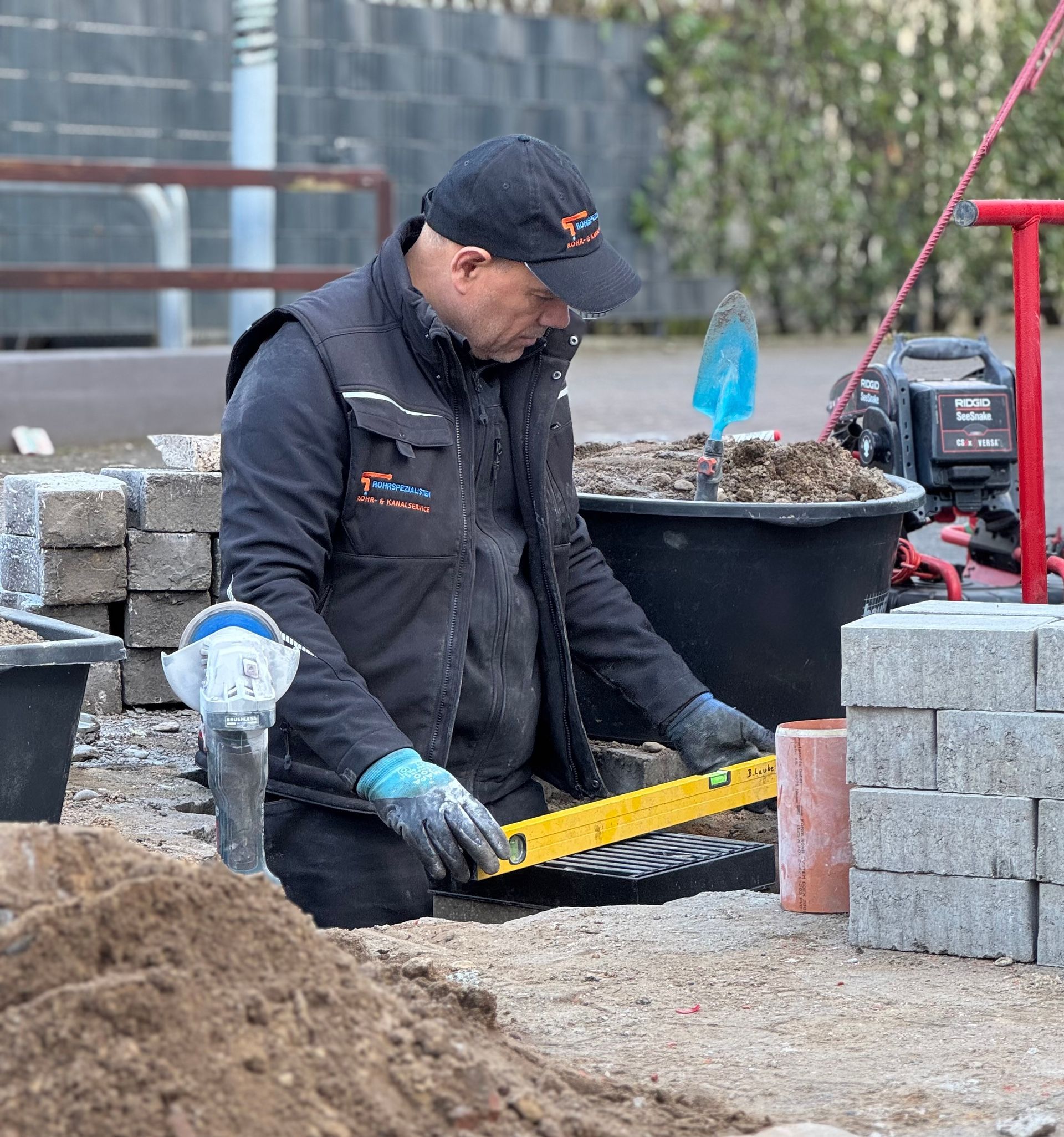 Ein Arbeiter in schwarzer Weste kniet an einem Gartenbeet und benutzt ein gelbes Werkzeug neben gestapelten Pflastersteinen und Erde.