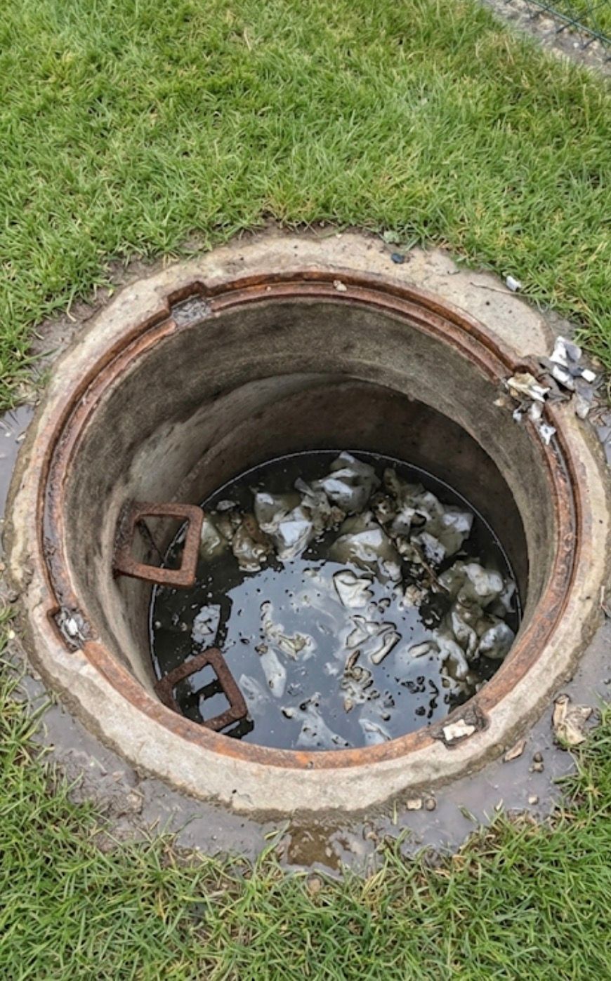 Offener Kanaldeckel mit stehendem Wasser und Schutt, umgeben von Gras und einem Betonrand
