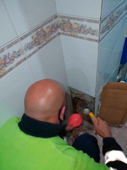 Hombre calvo con camisa verde, usando un martillo para romper baldosas en una esquina embaldosada.