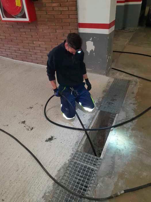 Un hombre vestido con ropa oscura y botas de seguridad se arrodilla para trabajar con una manguera en un desagüe de un garaje.