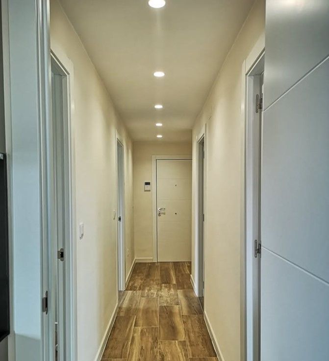 Un pasillo estrecho con paredes y puertas blancas, iluminación empotrada y suelos de imitación madera.
