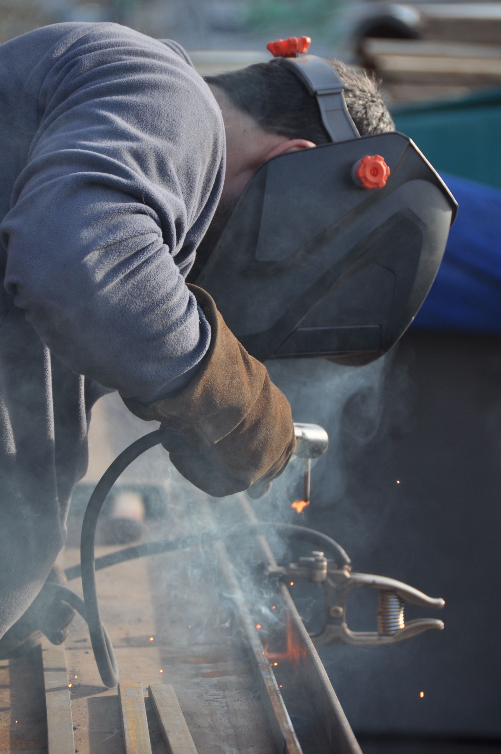 Un ouvrier soude du métal en extérieur, portant un masque de protection et des gants.