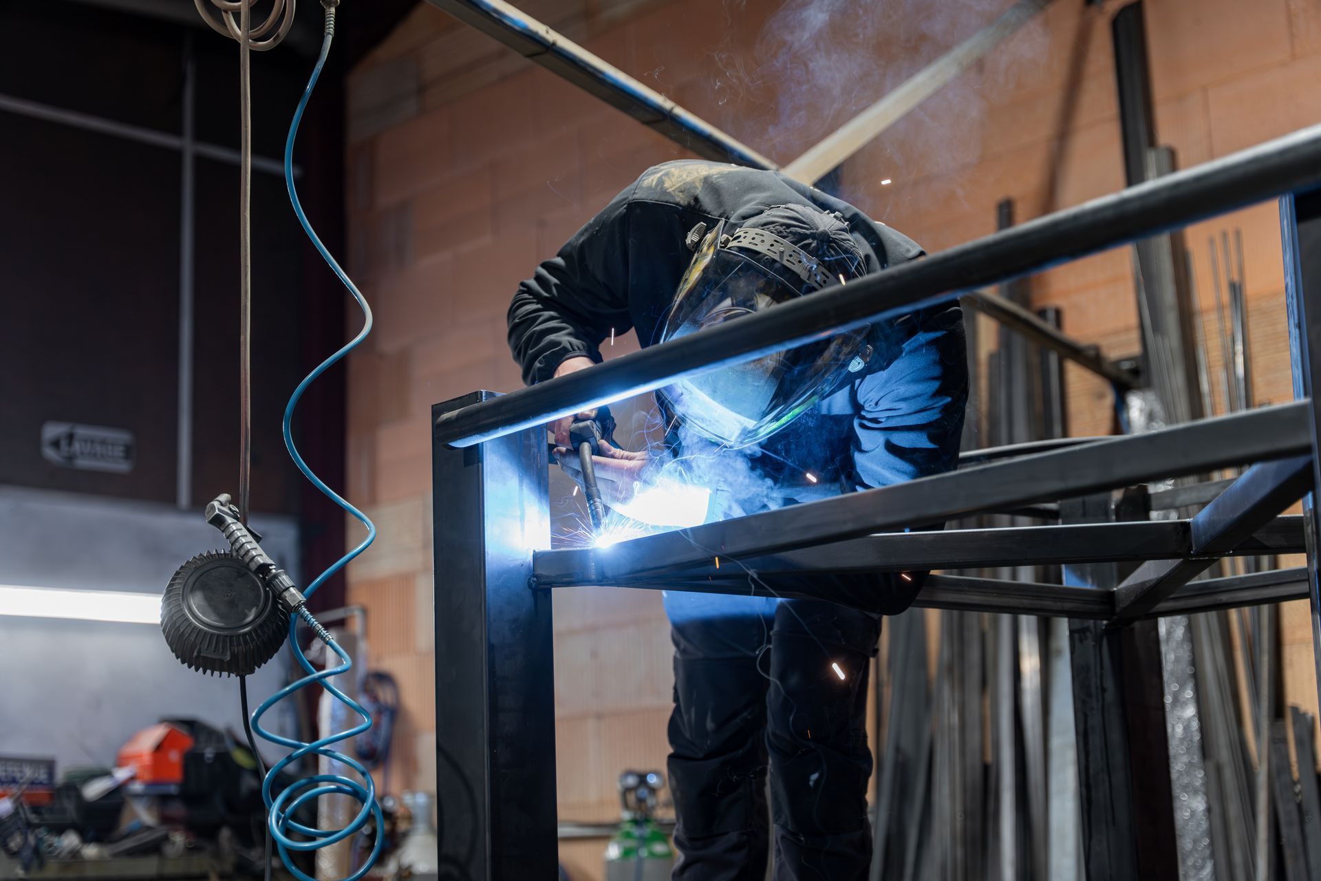 Un ouvrier soude un cadre métallique dans un atelier, avec des étincelles bleu vif et un câble suspendu à proximité.
