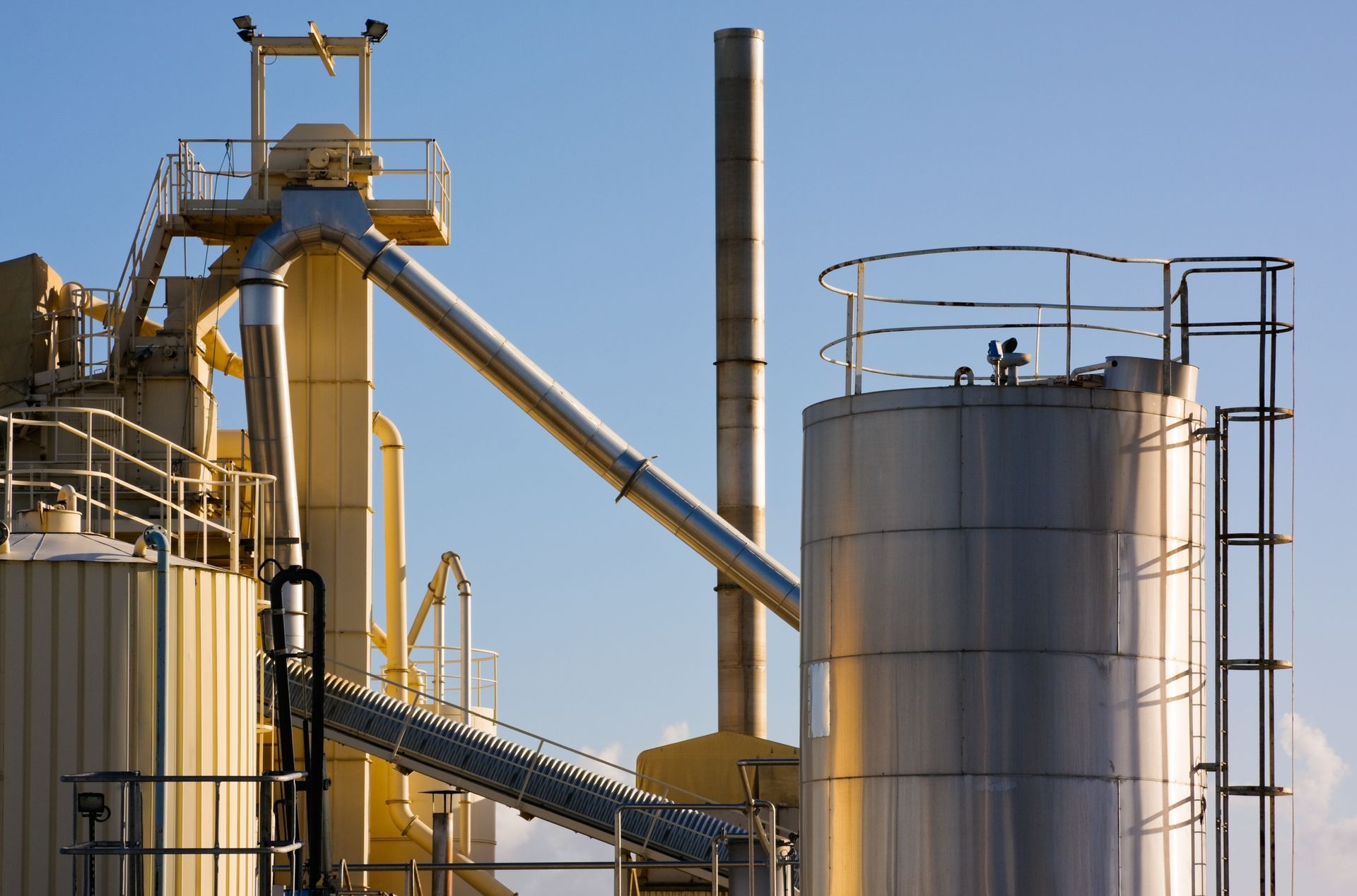 Site industriel avec des cuves de stockage argentées, des tuyauteries jaunes et de hautes cheminées sur fond de ciel dégagé.