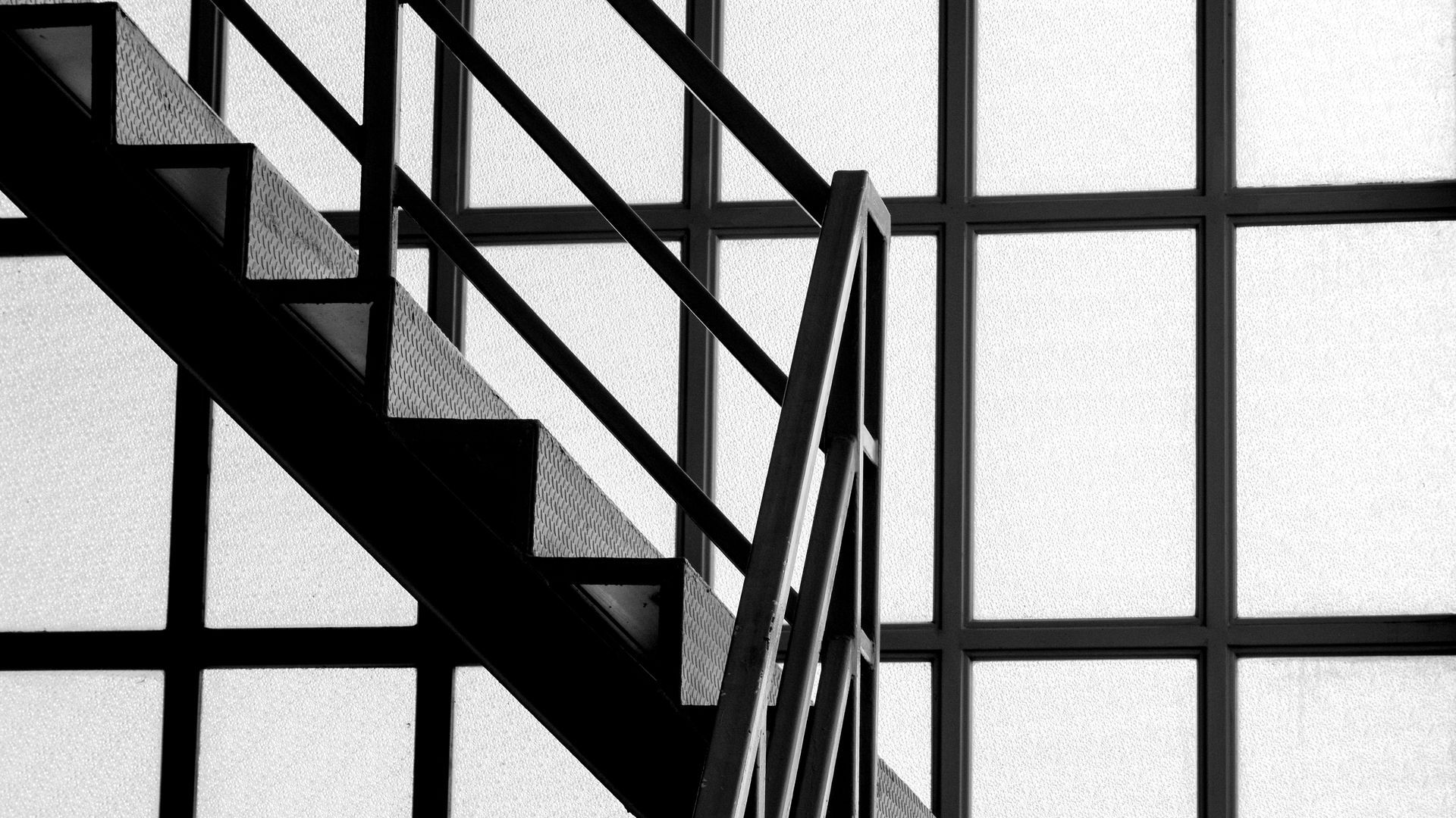 Escalier et rampes en métal noir se détachant sur une rangée de grandes fenêtres dépolies.
