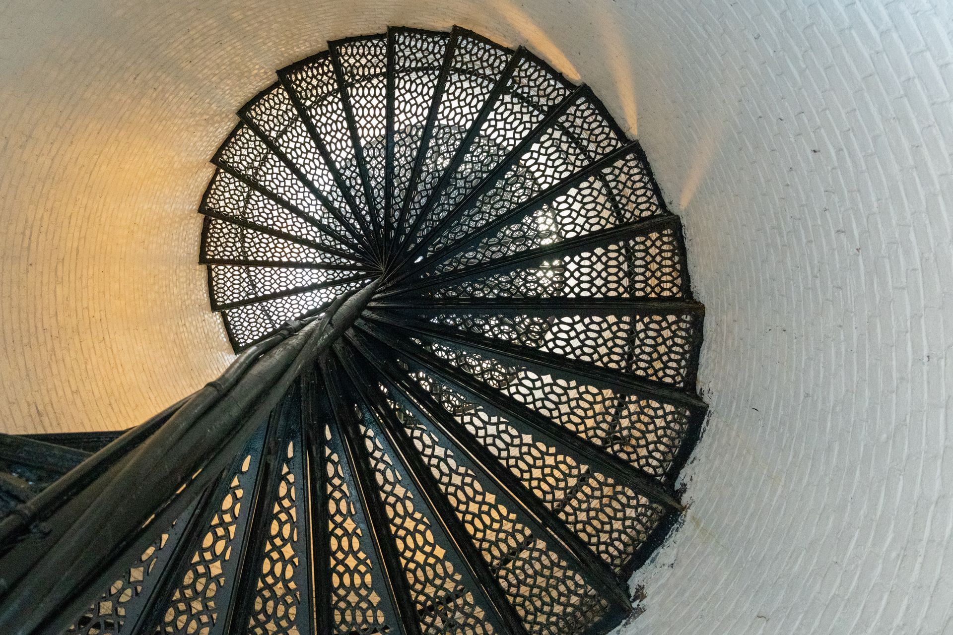 Escalier en colimaçon noir avec une rampe en métal ouvragée, contre un mur clair baigné d’une douce lumière chaude.