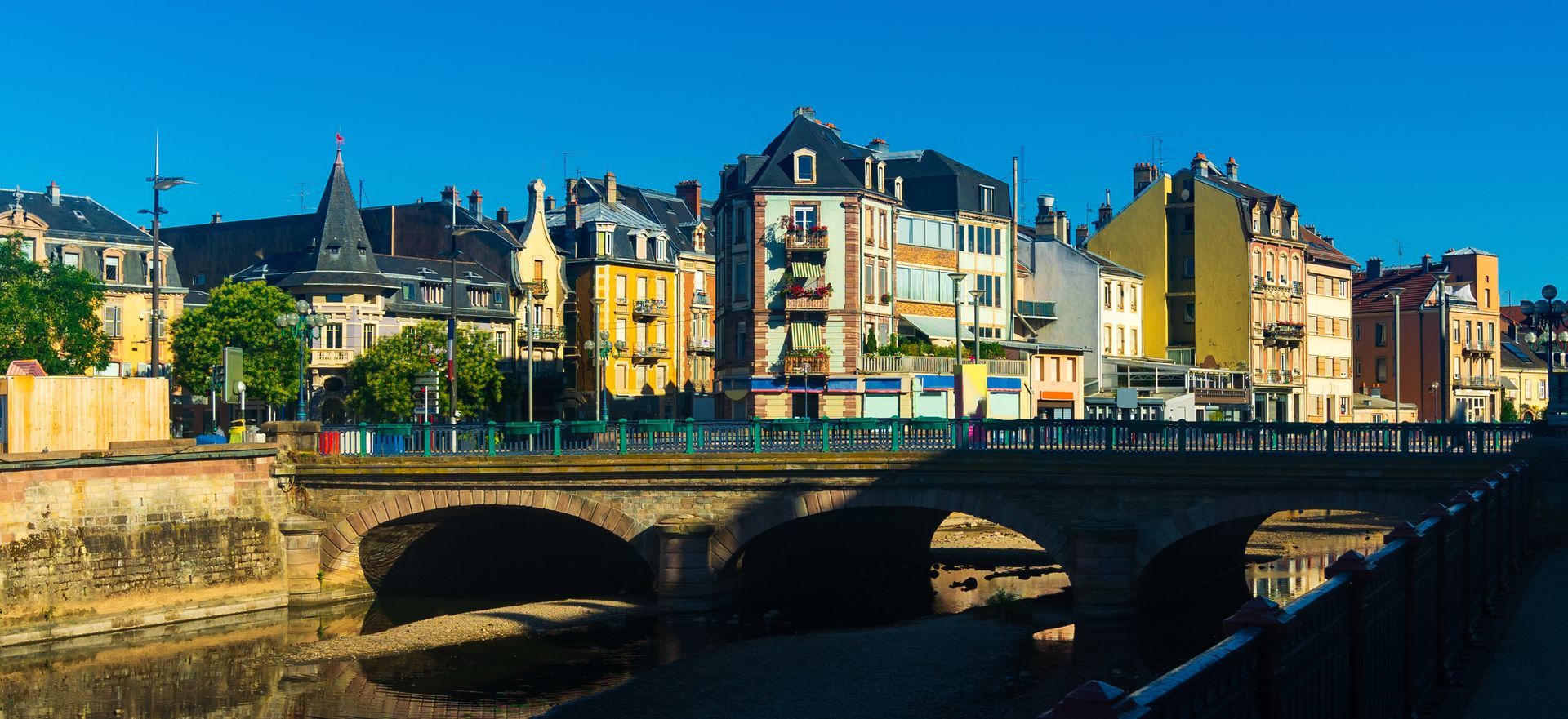 Des bâtiments colorés au bord de la rivière et un pont en pierre dans une ville européenne ensoleillée.