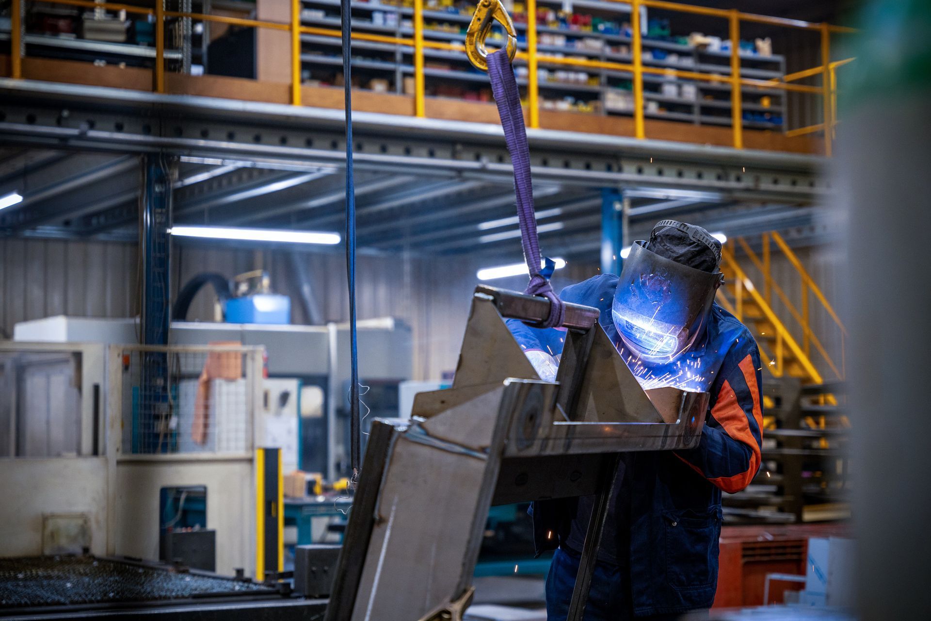 Un soudeur en tenue de sécurité orange fait jaillir des étincelles en travaillant le métal dans un atelier industriel.