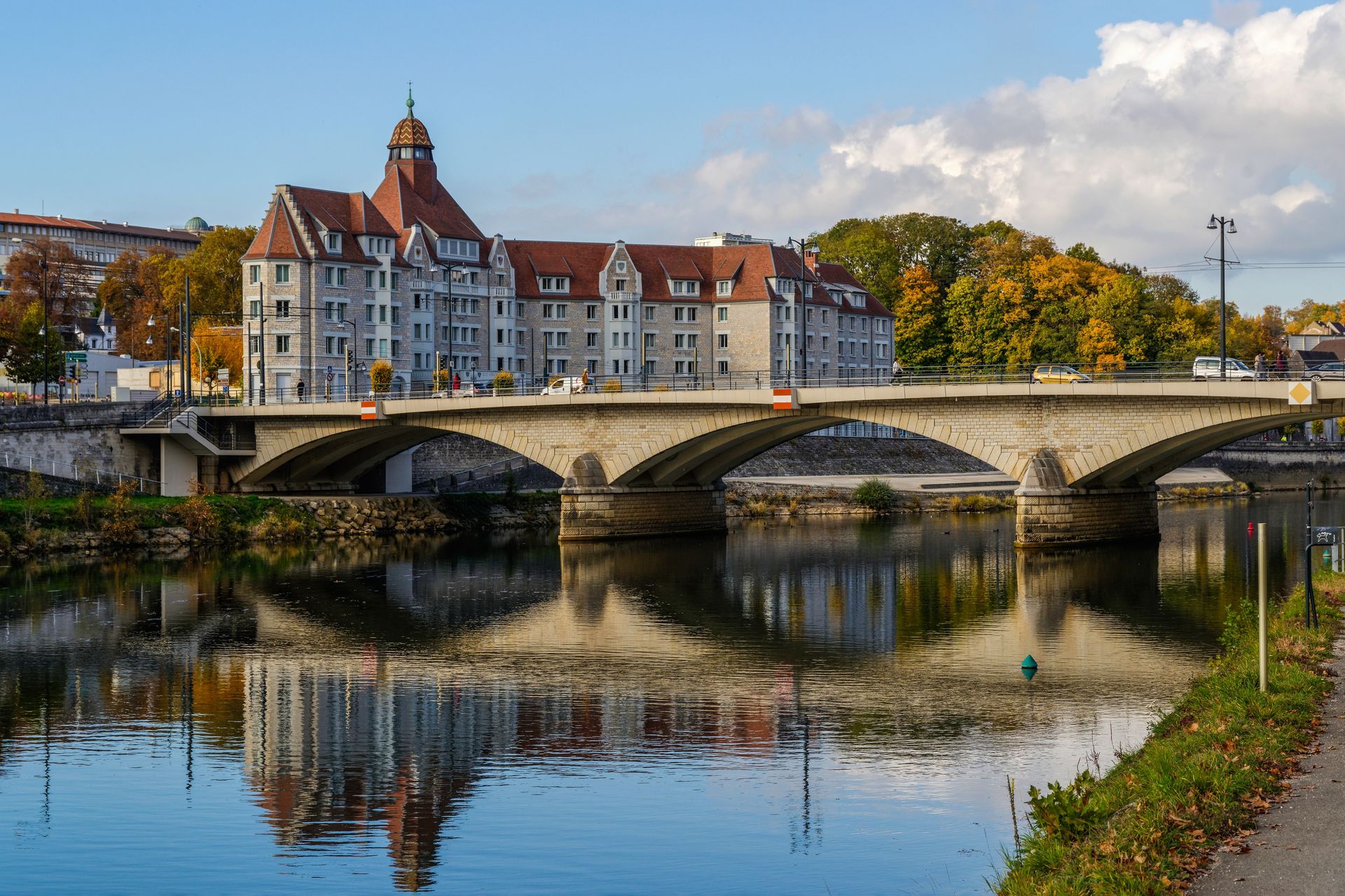 Pont de pierre au-dessus d’une rivière paisible, avec un grand bâtiment historique et des arbres d’automne qui se reflètent dans l’eau.