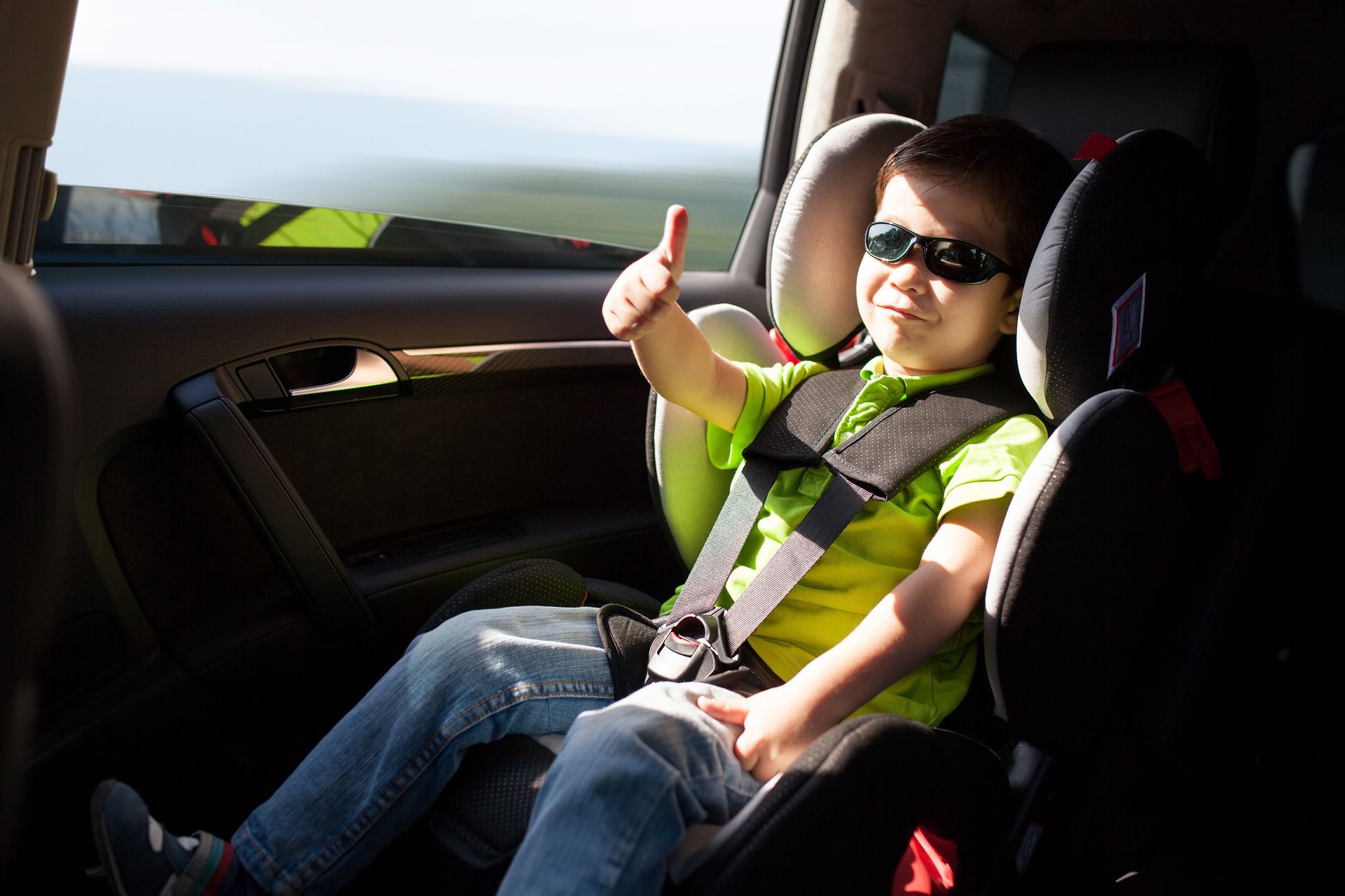 Un enfant portant des lunettes de soleil et un t-shirt vert est assis dans un siège auto et lève le pouce.