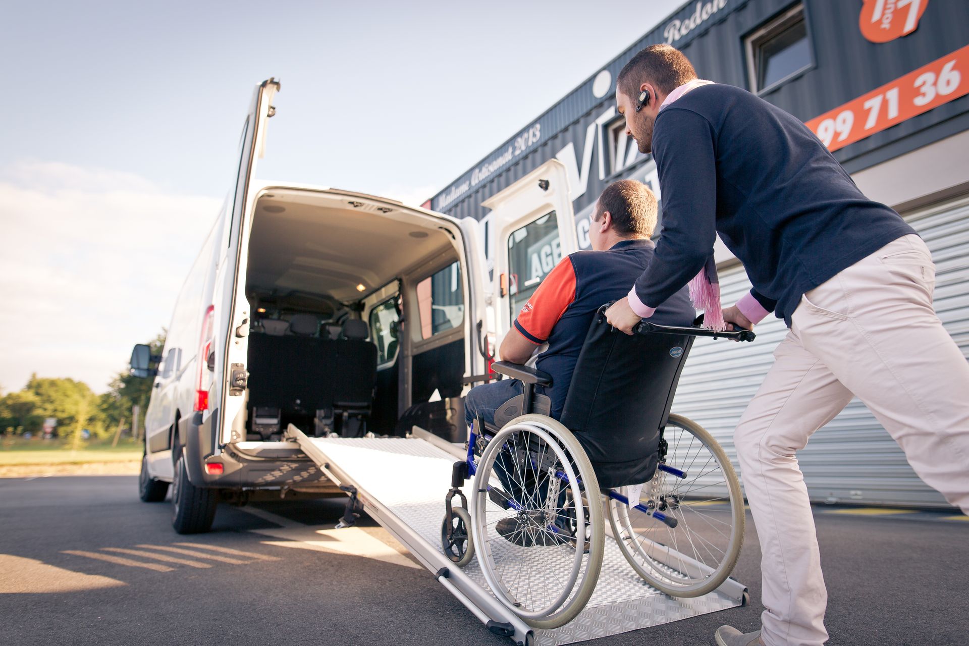 Une personne pousse un utilisateur de fauteuil roulant manuel sur une rampe pour le faire monter dans une camionnette blanche adaptée, garée devant un bâtiment commercial.