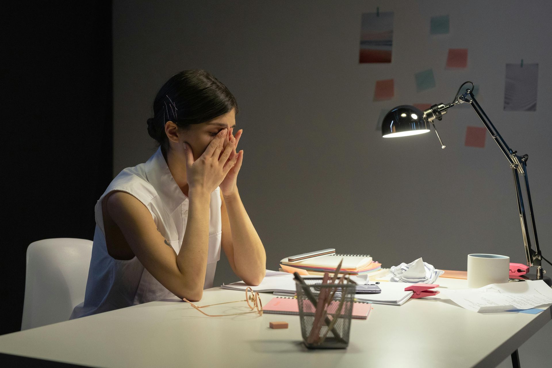 Mujer con las manos en la cara, luciendo estresada en un escritorio con papeles, una lámpara y útiles.