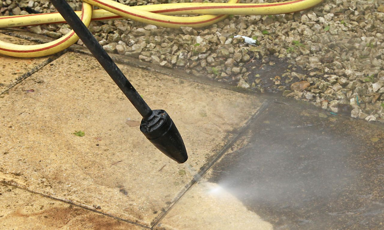 Nettoyeur haute pression pulvérisant des jets d'eau sur une terrasse en béton, en train d'enlever la saleté ; on aperçoit l'eau et le tuyau jaune.